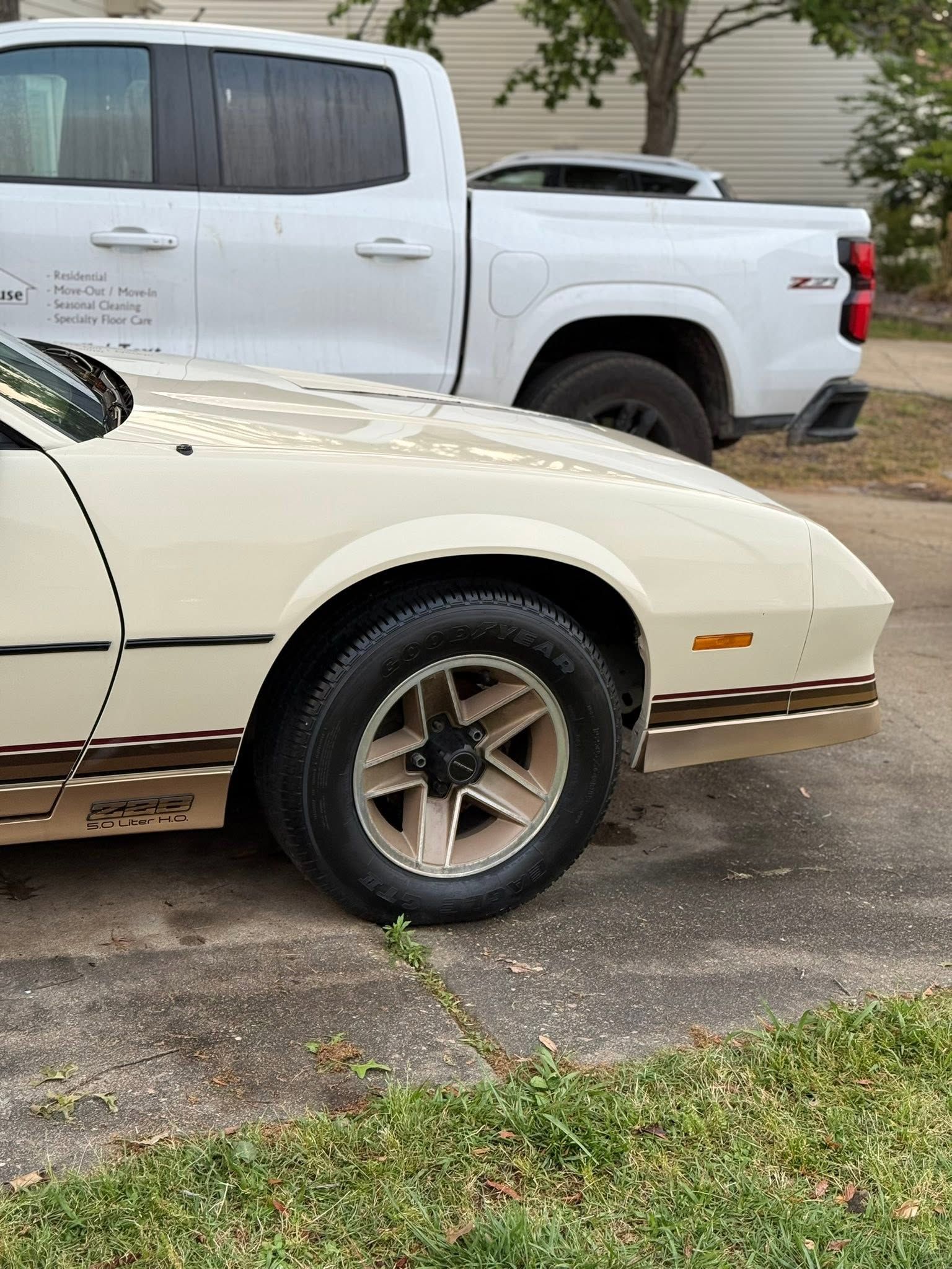 Cream vintage car parked beside a white pickup on a driveway near grass and trees.