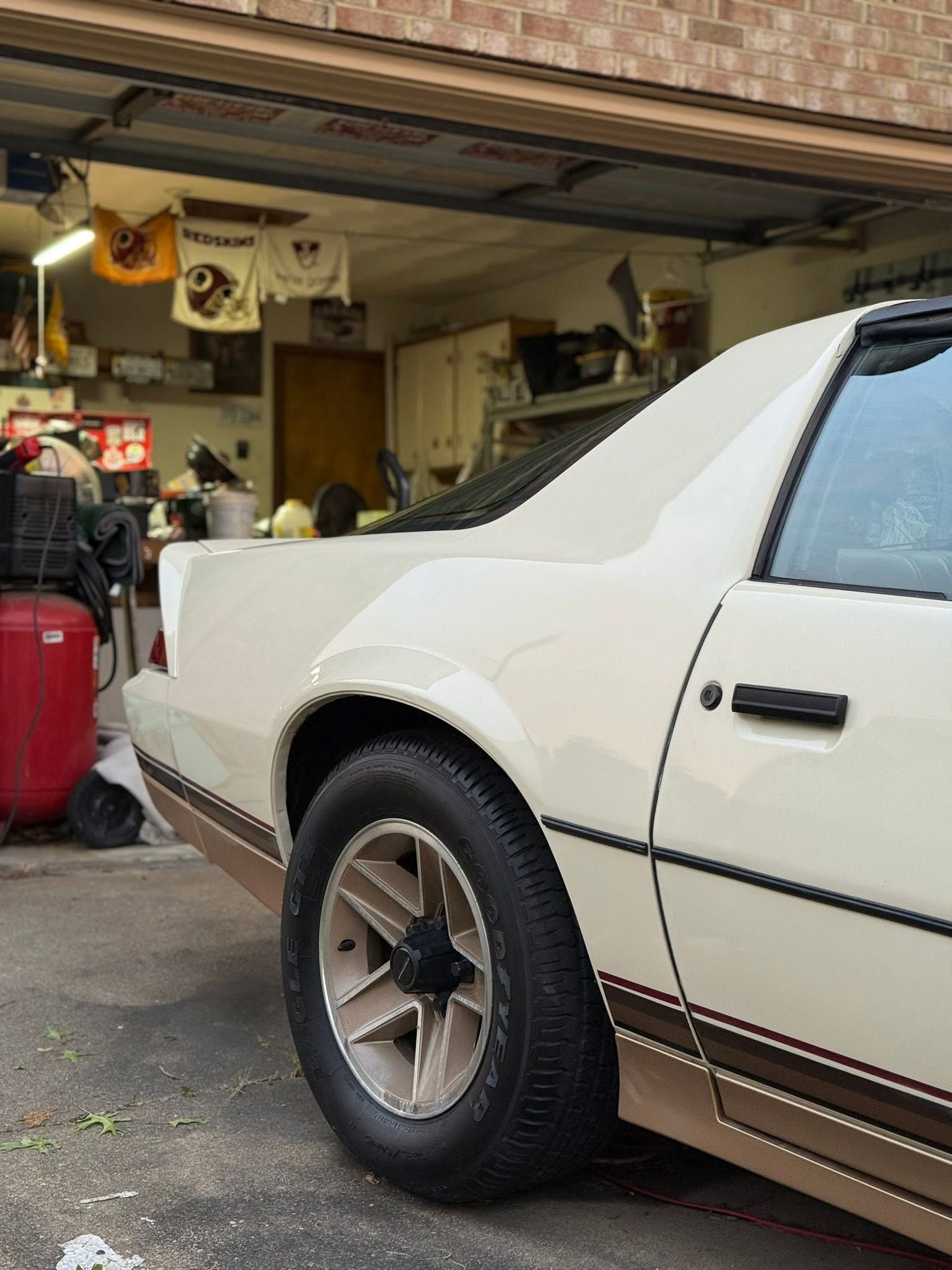 White vintage coupe parked in a cluttered garage, rear side view with a black wheel.