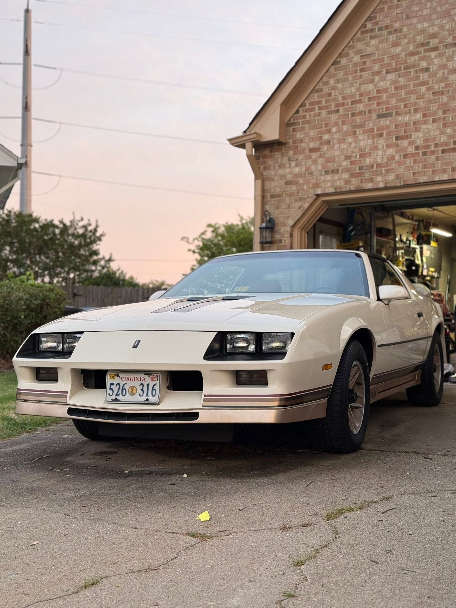White sports car partially out of a garage at dusk, front bumper damaged, parked on a driveway