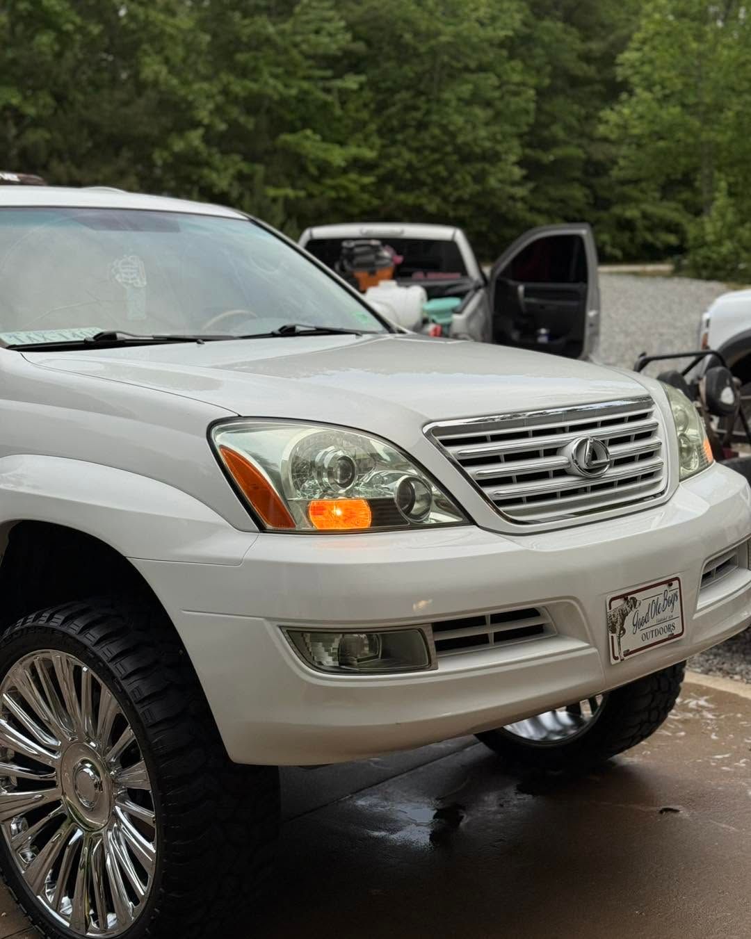 Silver SUV parked outside with open door; person loading items near another vehicle in a wooded area.