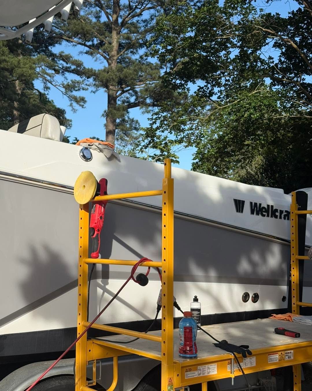 Yellow scaffolding beside a white Welktra trailer under trees, with tools on the platform