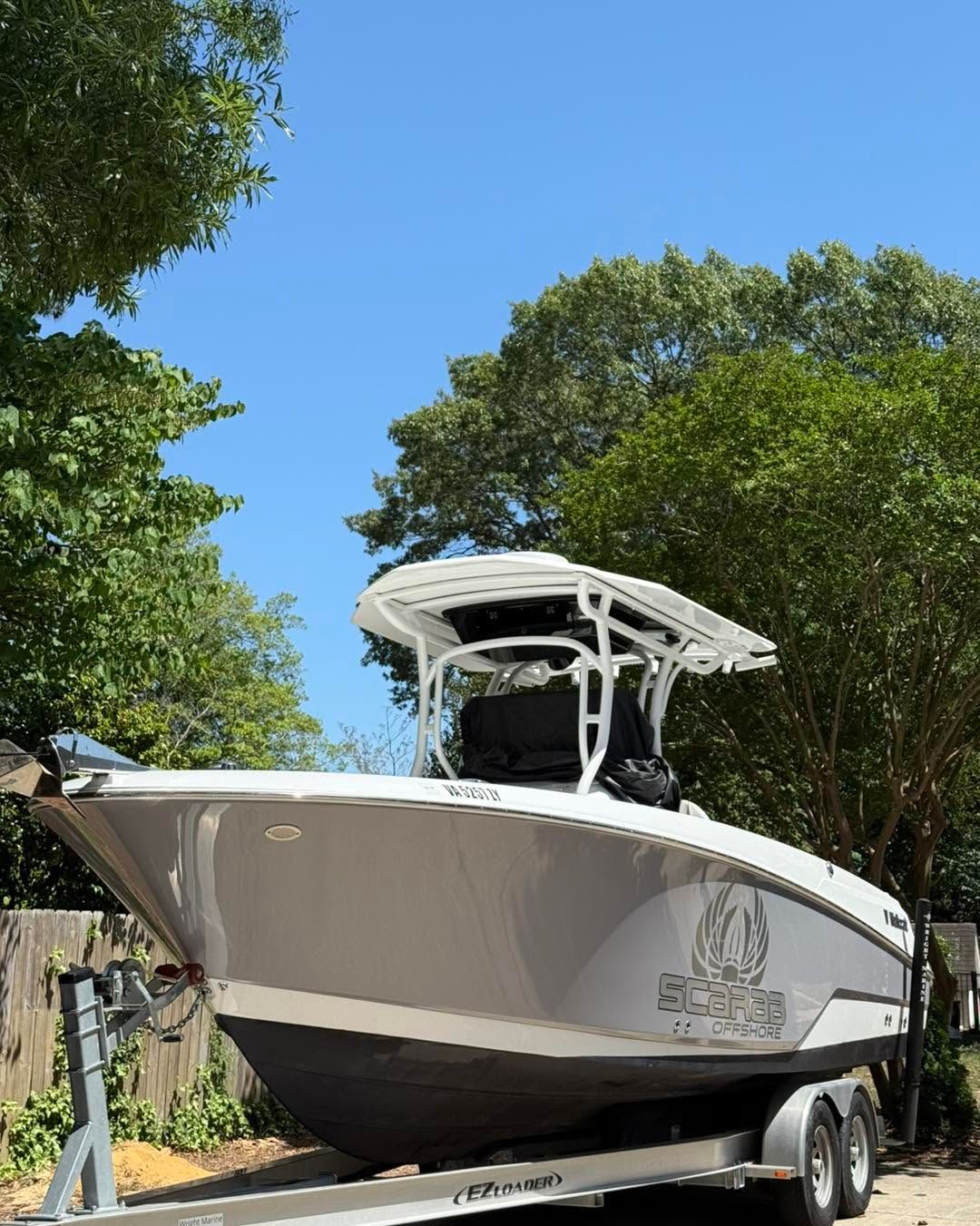 White fishing boat on a trailer, parked outdoors under trees against a blue sky.