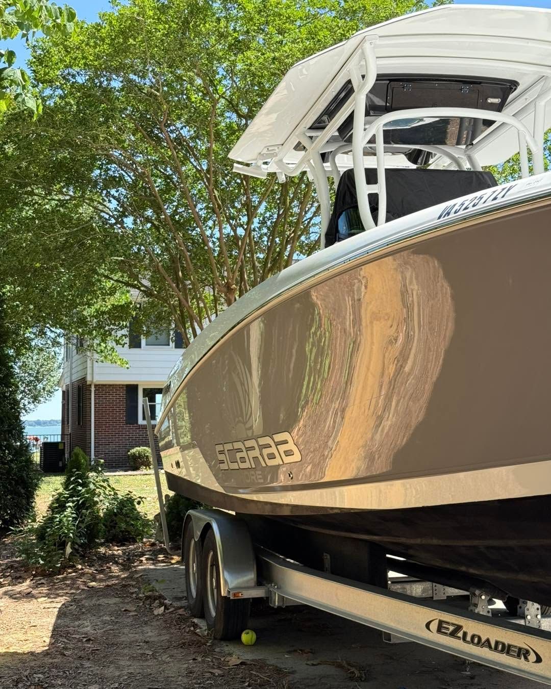 White boat on trailer parked under trees beside a house, with a visible curved hull and canopy.
