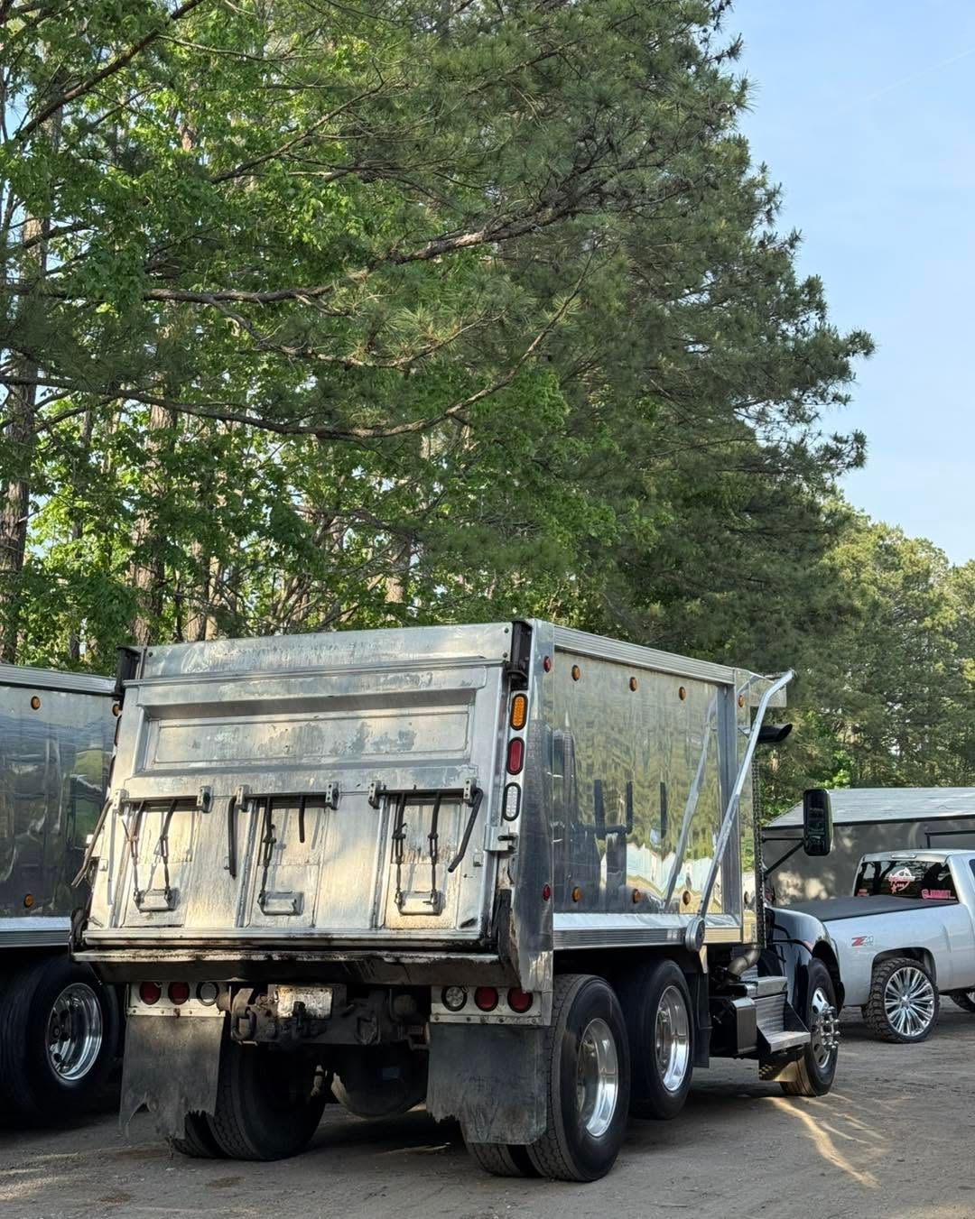 Silver dump truck parked on a roadside under tall green trees, with cars and a white SUV nearby.
