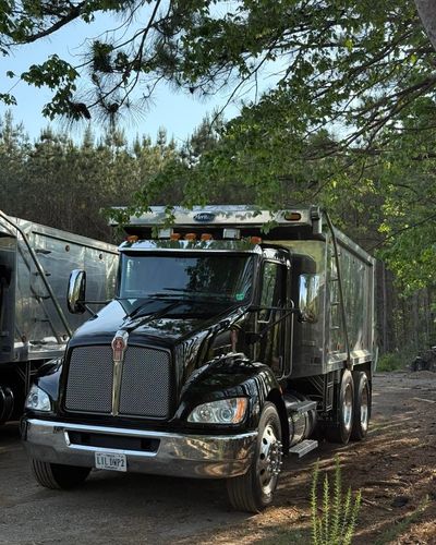 Black semi-truck with silver trailer parked on a wooded dirt road