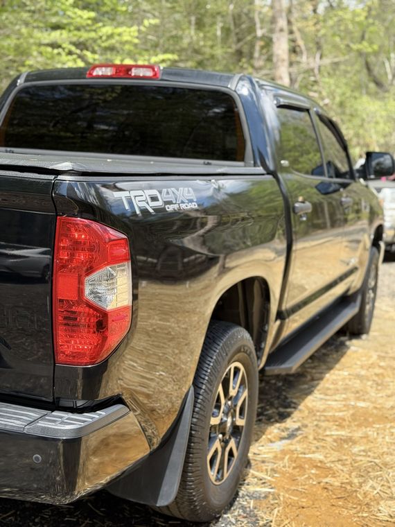 Black pickup truck parked on a dirt path in a wooded area, rear and side view.