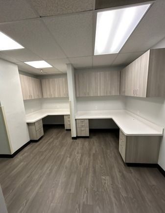 Office space with two L-shaped desks, cabinets, and wood-look flooring. Light beige cabinetry.