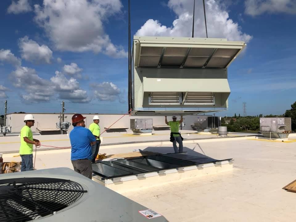 Workers installing HVAC unit on a rooftop. Crane lifting a large, rectangular unit. Sunny, blue sky.