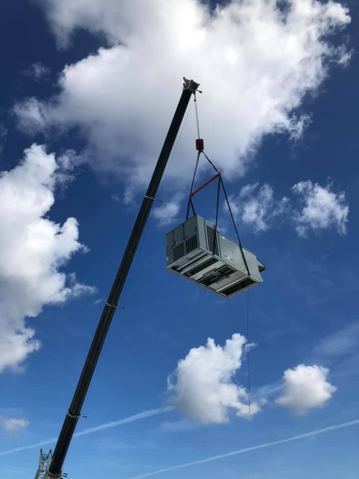 A crane lifting a large HVAC unit against a blue sky with fluffy clouds.