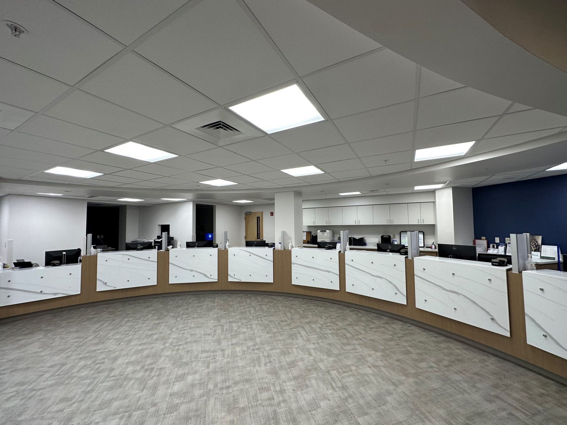 Circular office reception area with white desks, computers, and gray carpet. Fluorescent lights illuminate the ceiling.