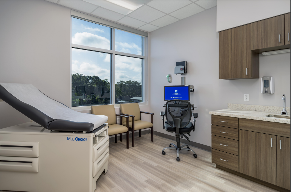 Medical examination room with exam table, chairs, cabinets, and a window overlooking greenery.