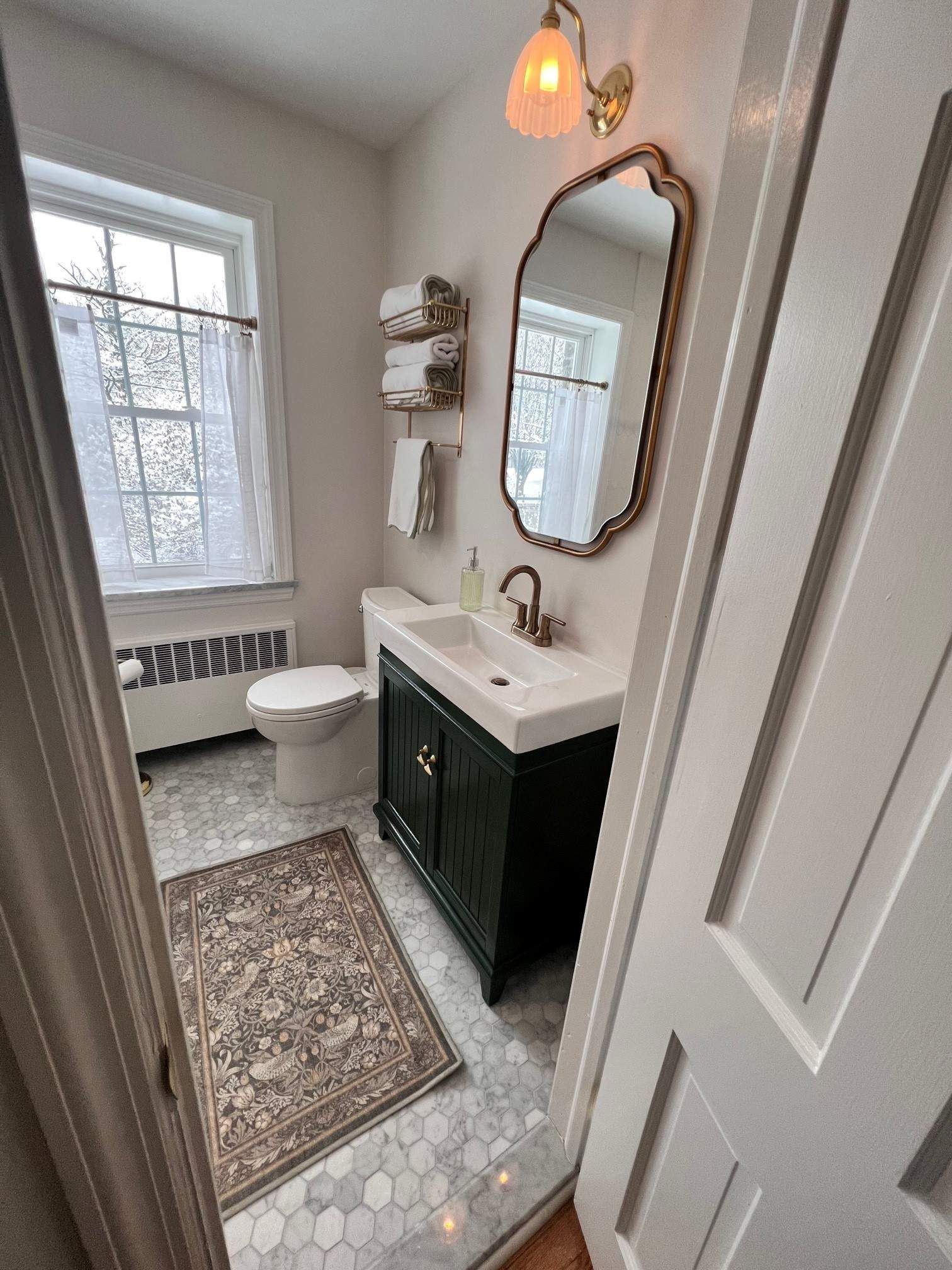 Small bathroom with green vanity, patterned rug, white toilet, and gold mirror.