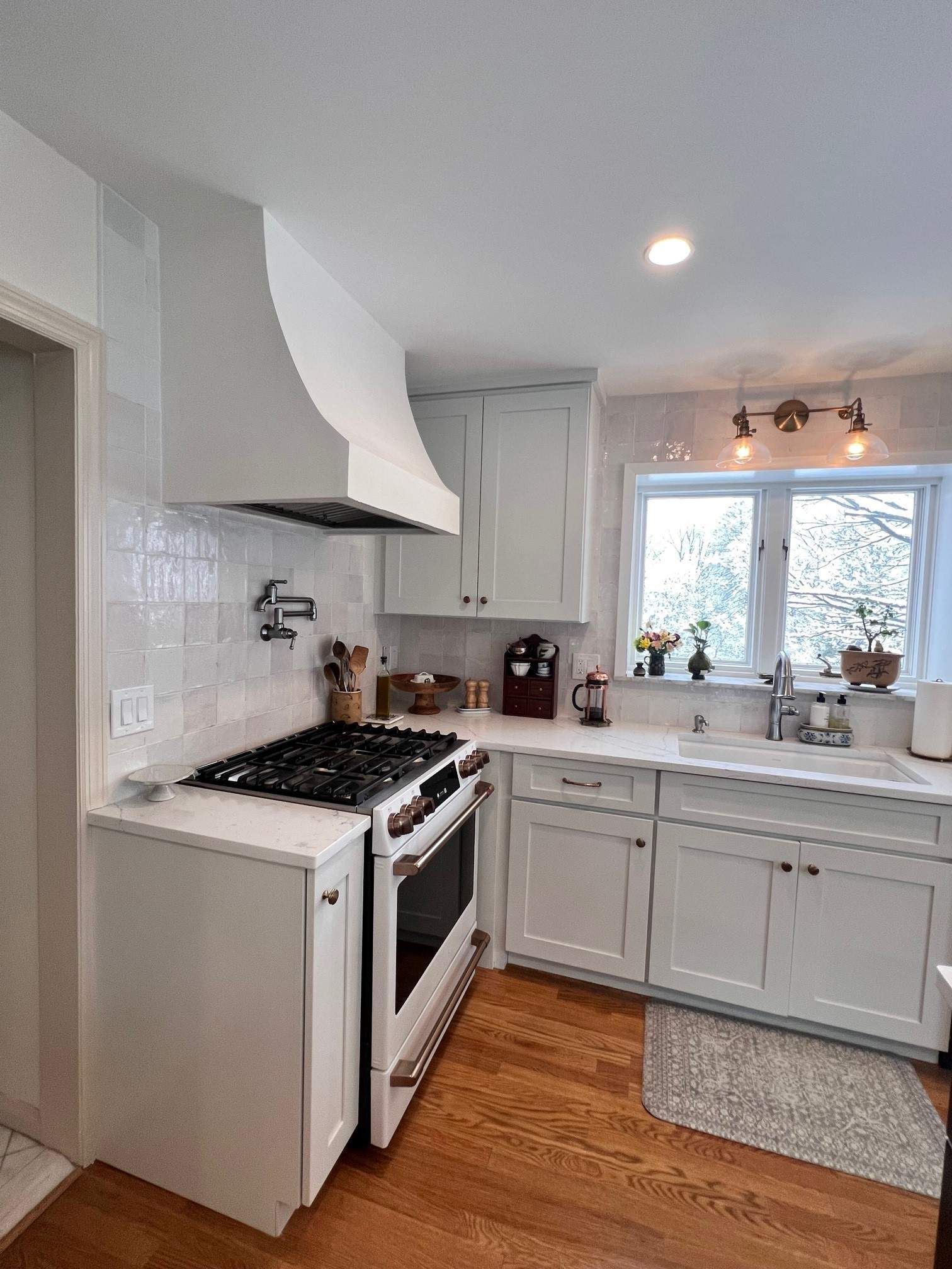White kitchen with stove, cabinets, and window. Wood floors, tile backsplash, and range hood.