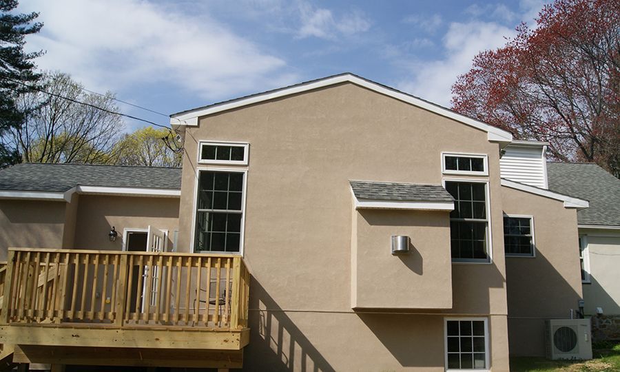 Back view of a light brown house with a wooden deck, several windows, and a blue sky.