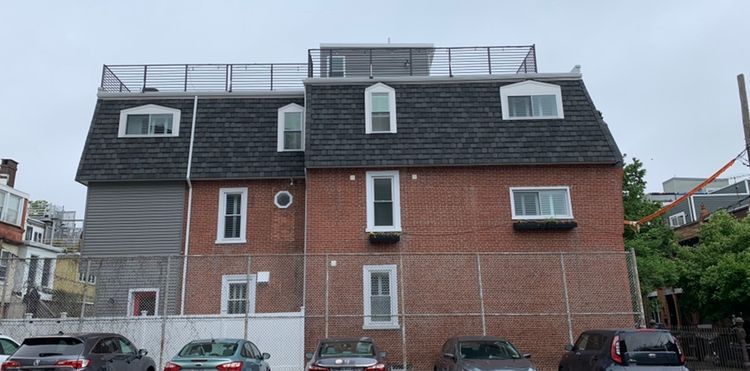 Brick building with dark roof, windows, and rooftop structure. Cars parked in front of a white fence.