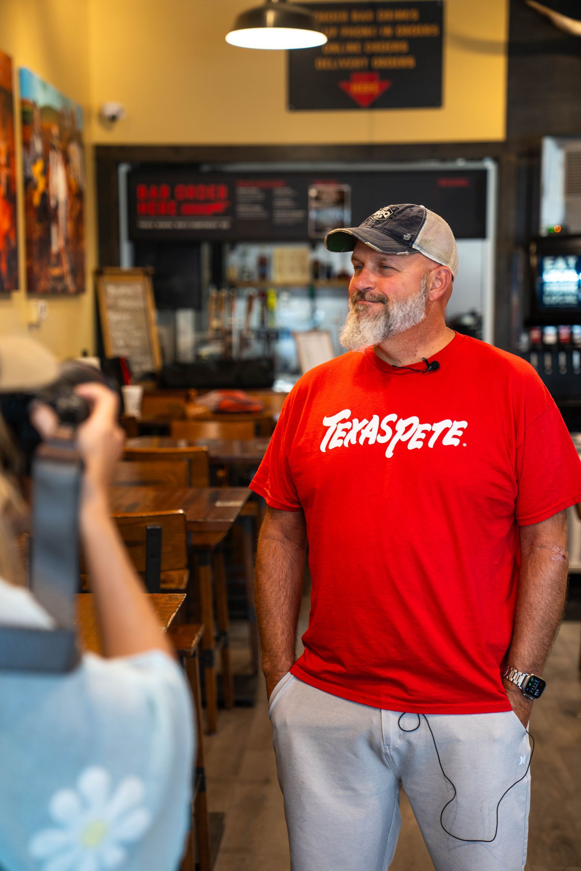 Man in red T-shirt and gray pants standing indoors, smiling near a counter in a café or shop.