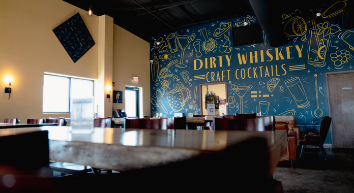 Restaurant interior with large “Dirty Whiskey Craft Cocktails” mural and warm booth seating