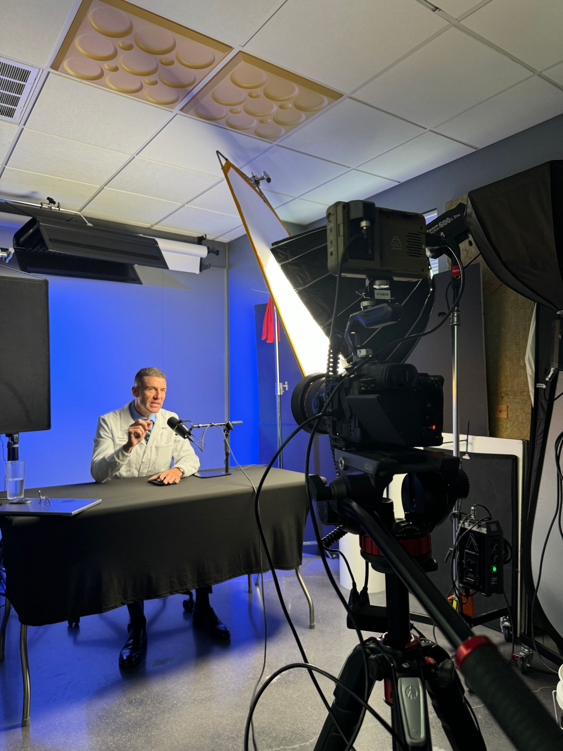 A man sits at a desk in a studio, speaking on camera under blue lighting with a large camera in the foreground.