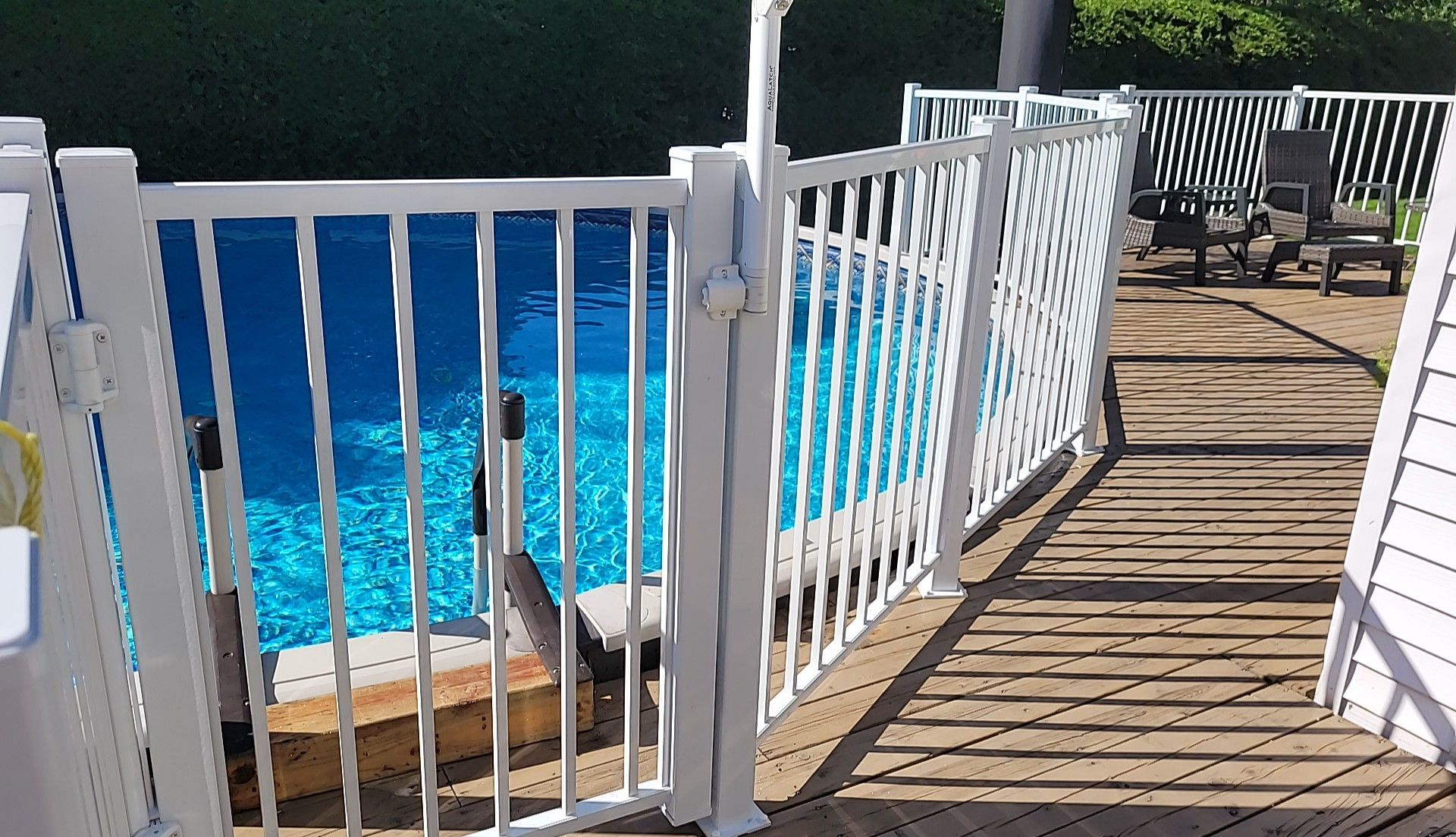 Une clôture blanche entoure une piscine à l'eau bleue. On aperçoit une terrasse en bois et de la verdure.
