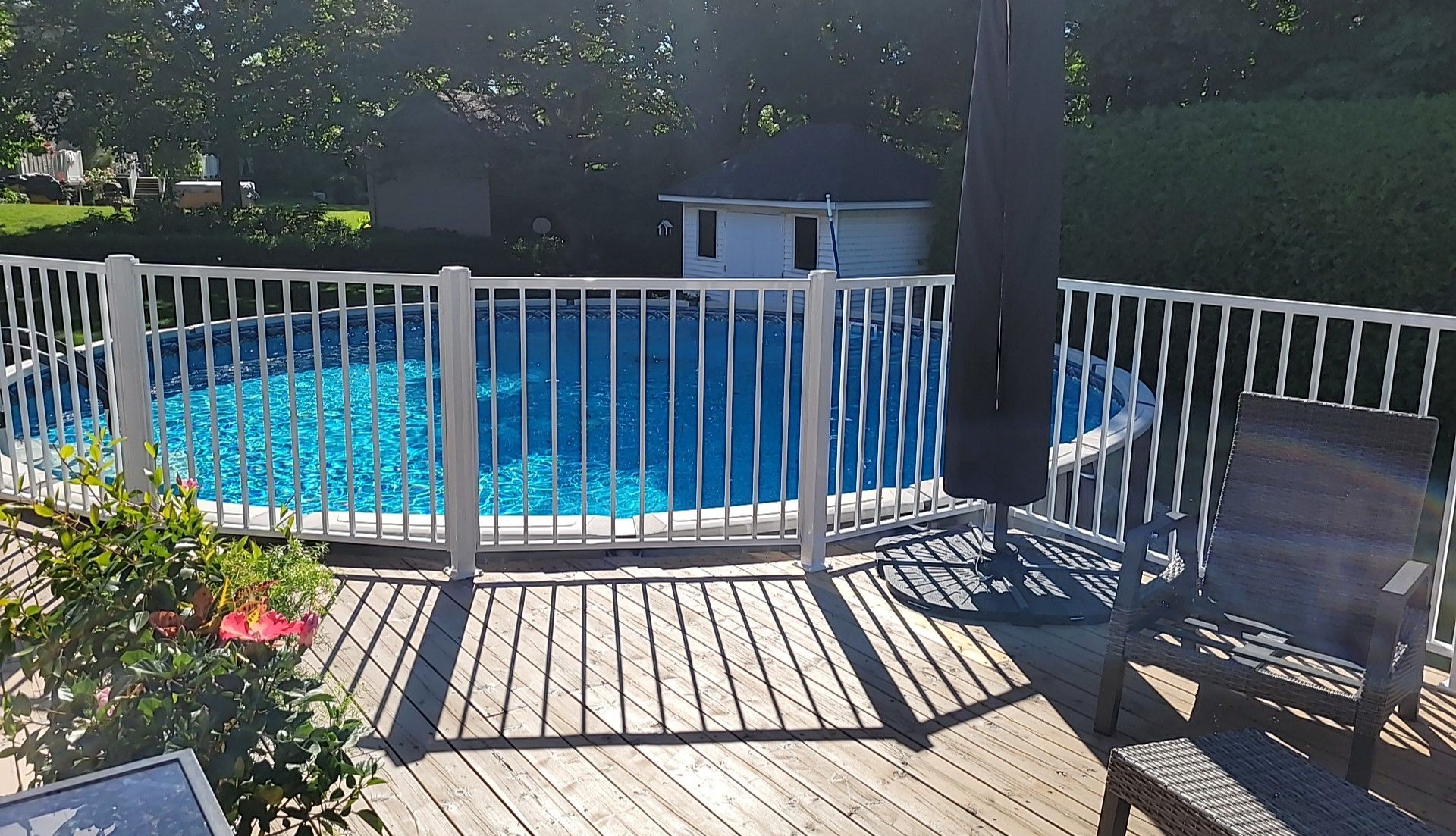 Piscine hors sol entourée d'une clôture blanche sur une terrasse en bois, avec parasol et chaise noirs.