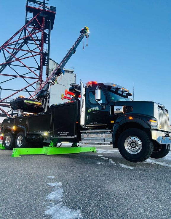 A black Western Star heavy-duty tow truck with an extended crane, parked outdoors on a snowy lot with a red tower behind.