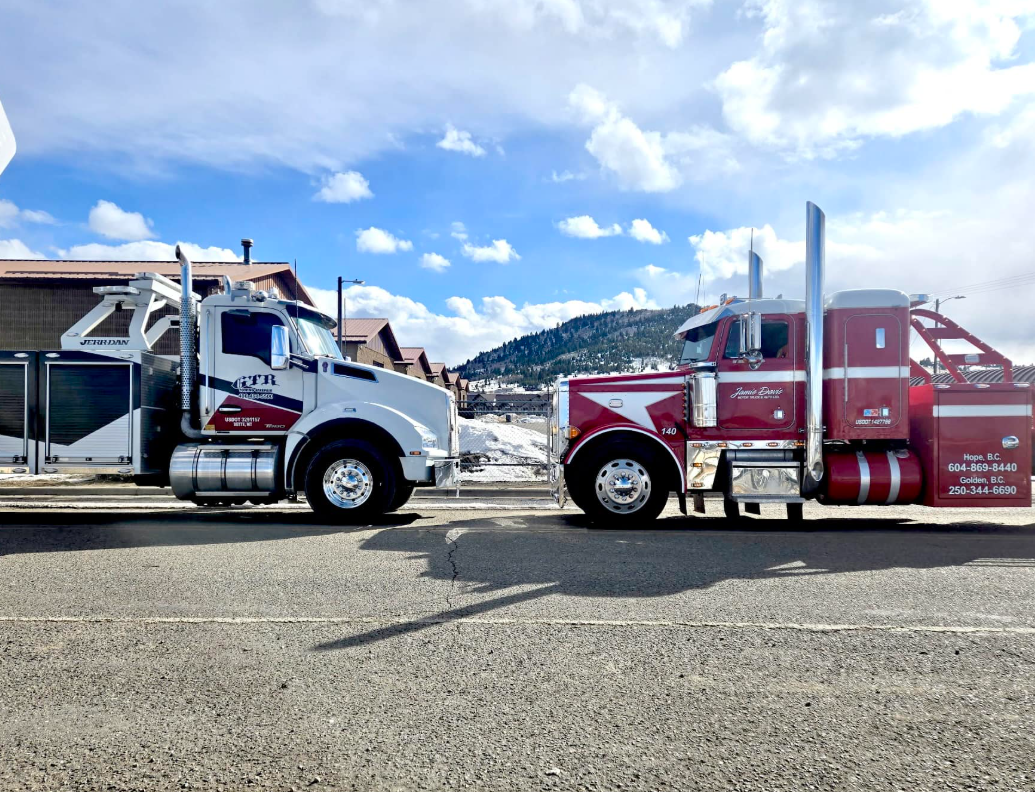 A white and a red semi-truck tow truck are parked side-by-side in a gravel lot with mountains in the background.