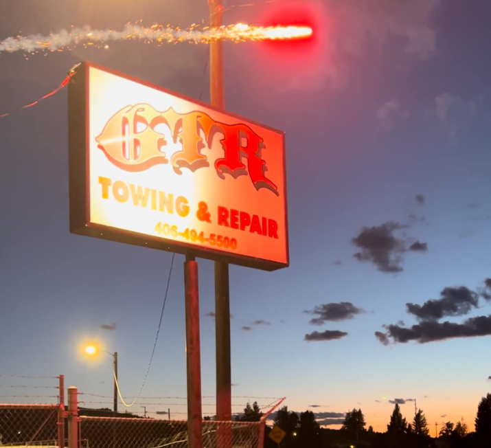 A sign for GTR Towing & Repair stands against a twilight sky with a bright red light flare trailing through the air.