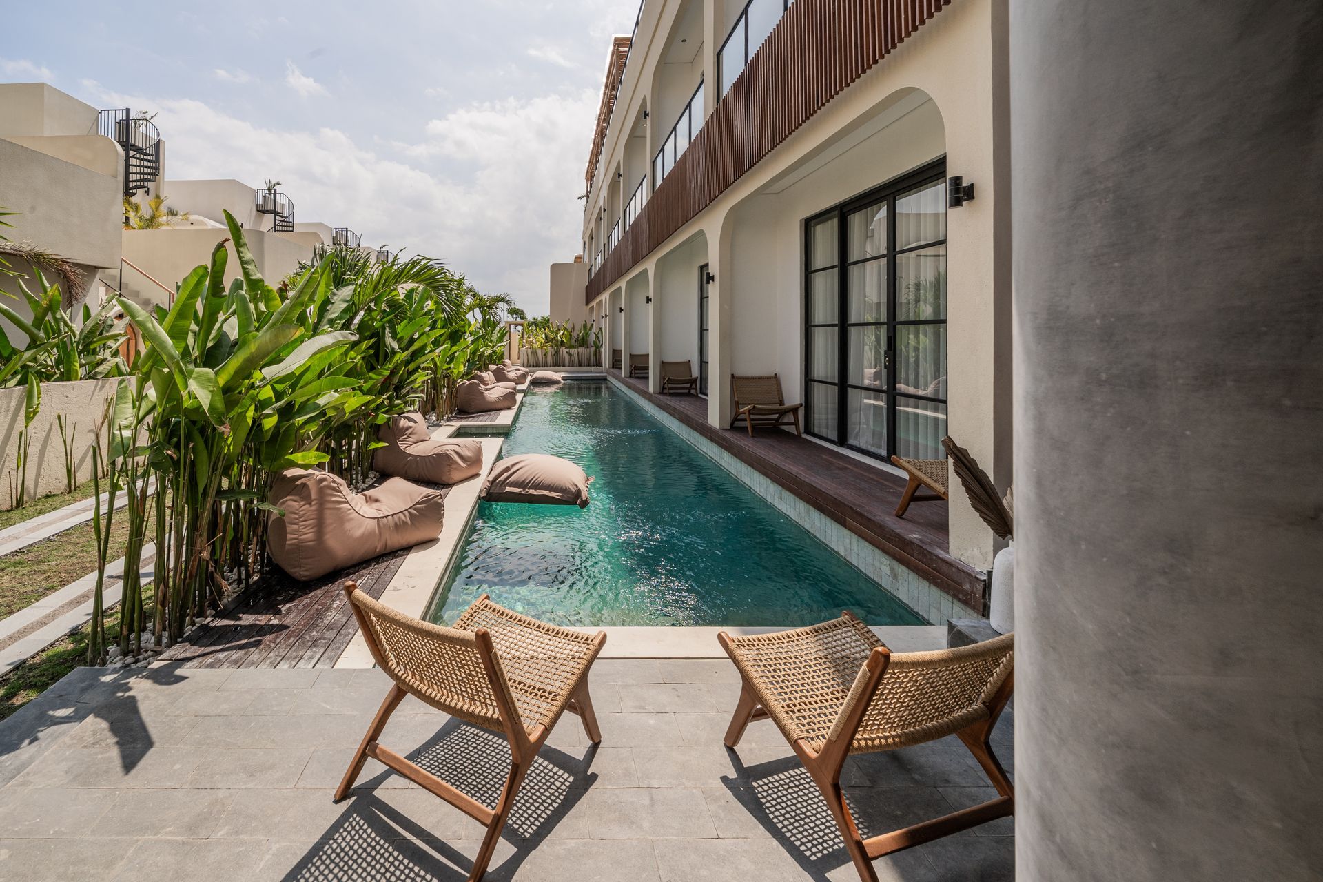 Modern patio with a small turquoise pool, white walls, stone backdrop, and lush tropical plants.