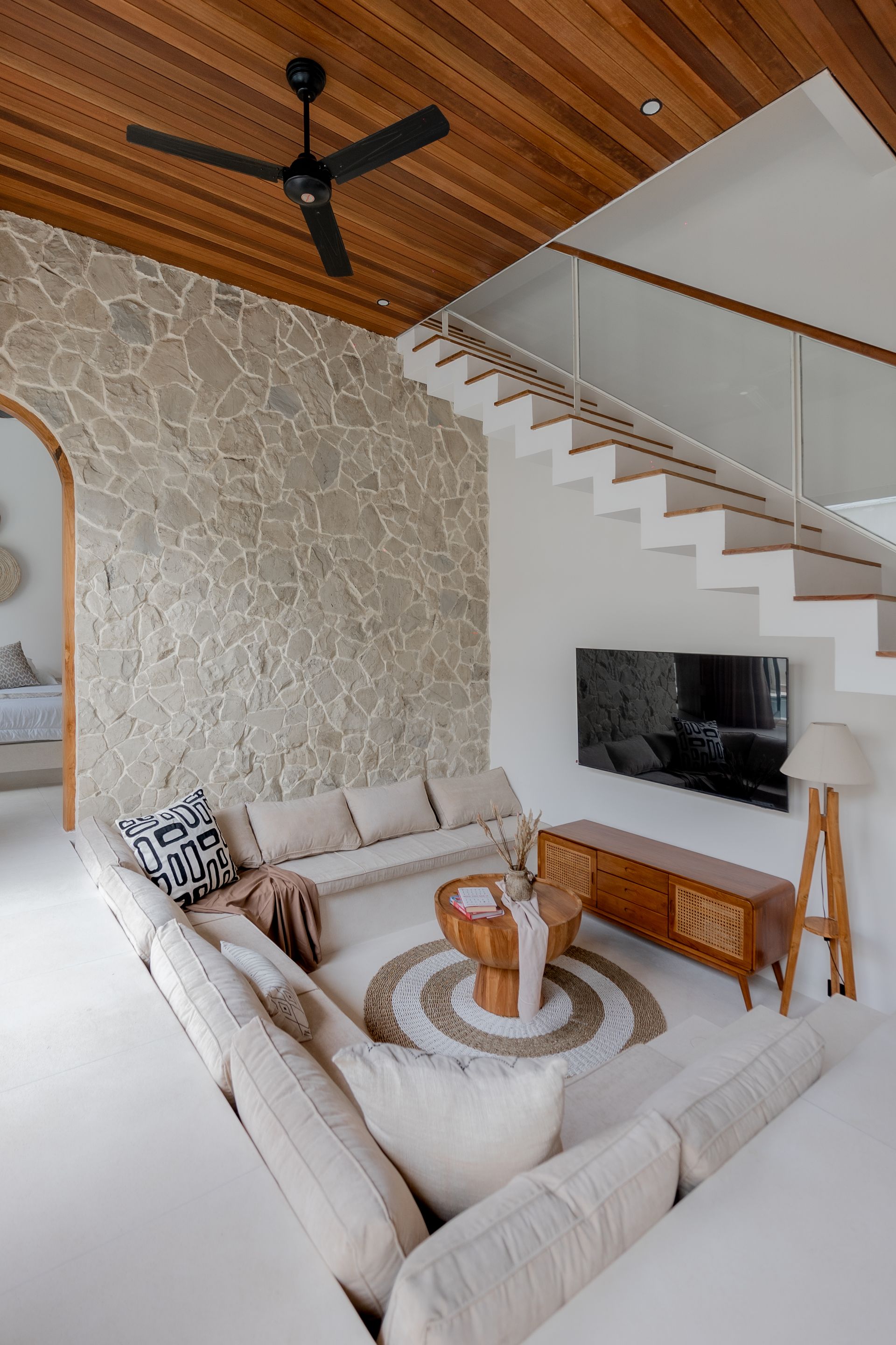 Cozy living room with white sectional, stone accent wall, wood ceiling, stairs, and black ceiling fan