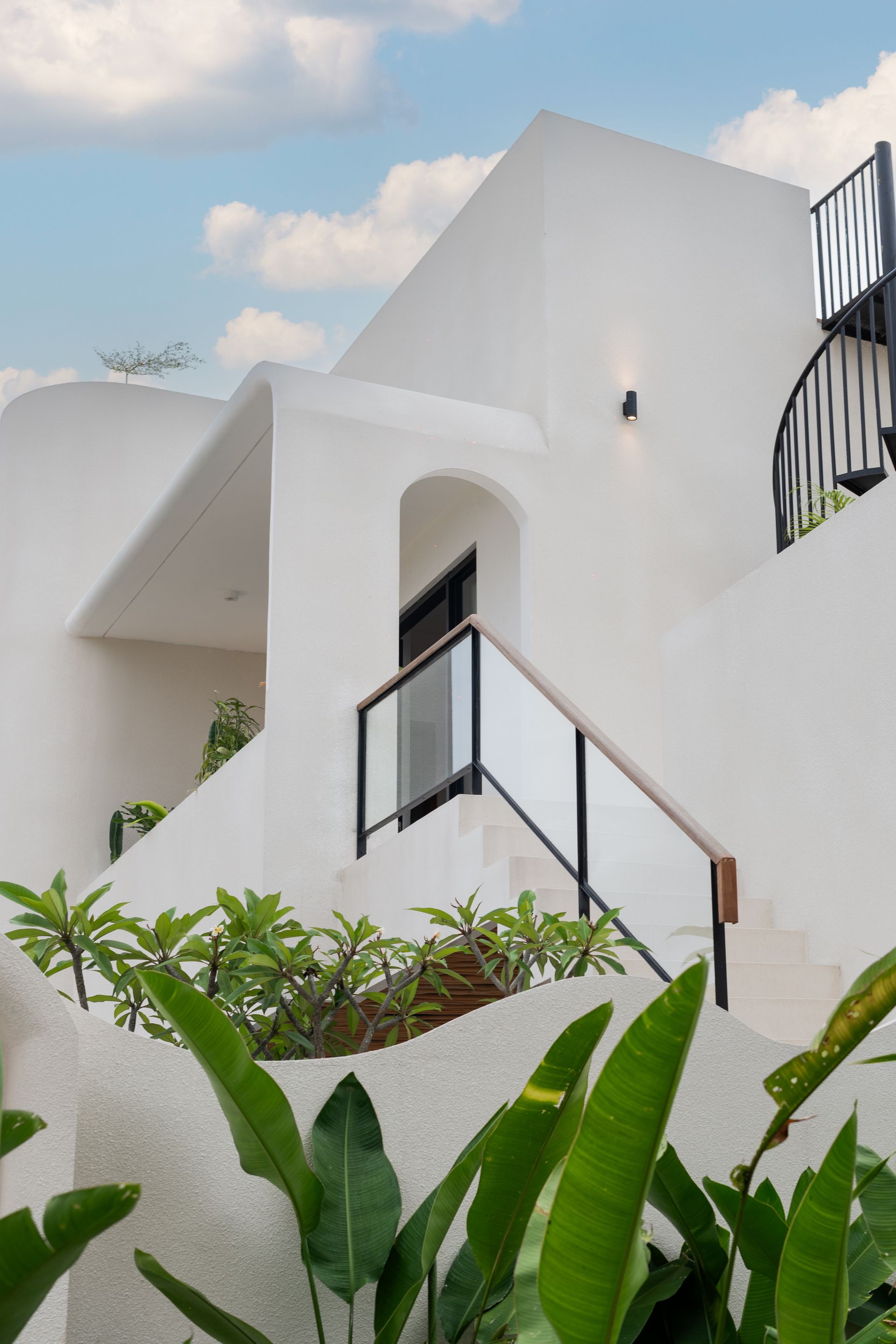 White modern building with curved stairs, black railings, and lush green plants in front under a blue sky