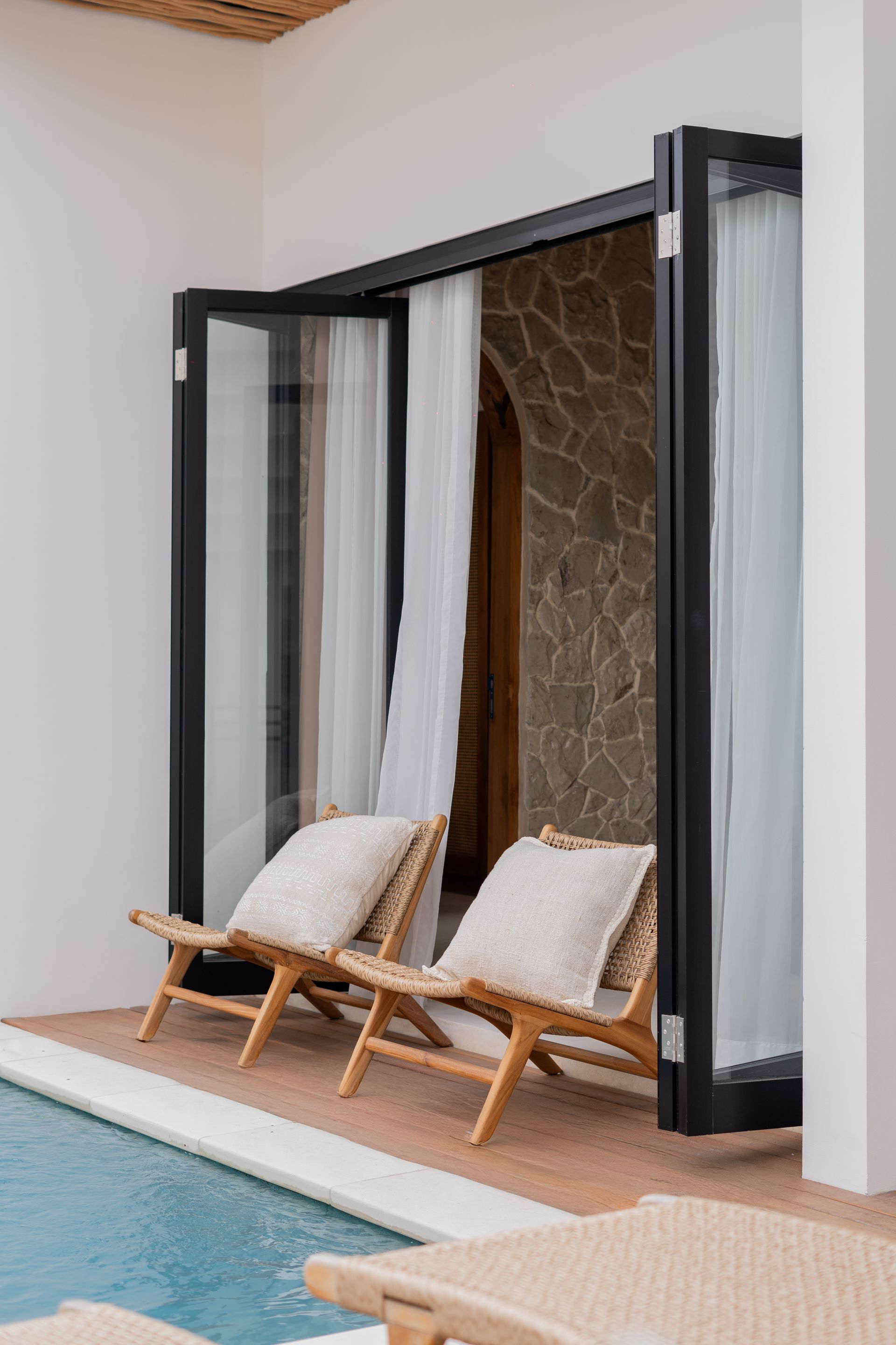 Poolside patio with open black-framed doors and two beige lounge chairs