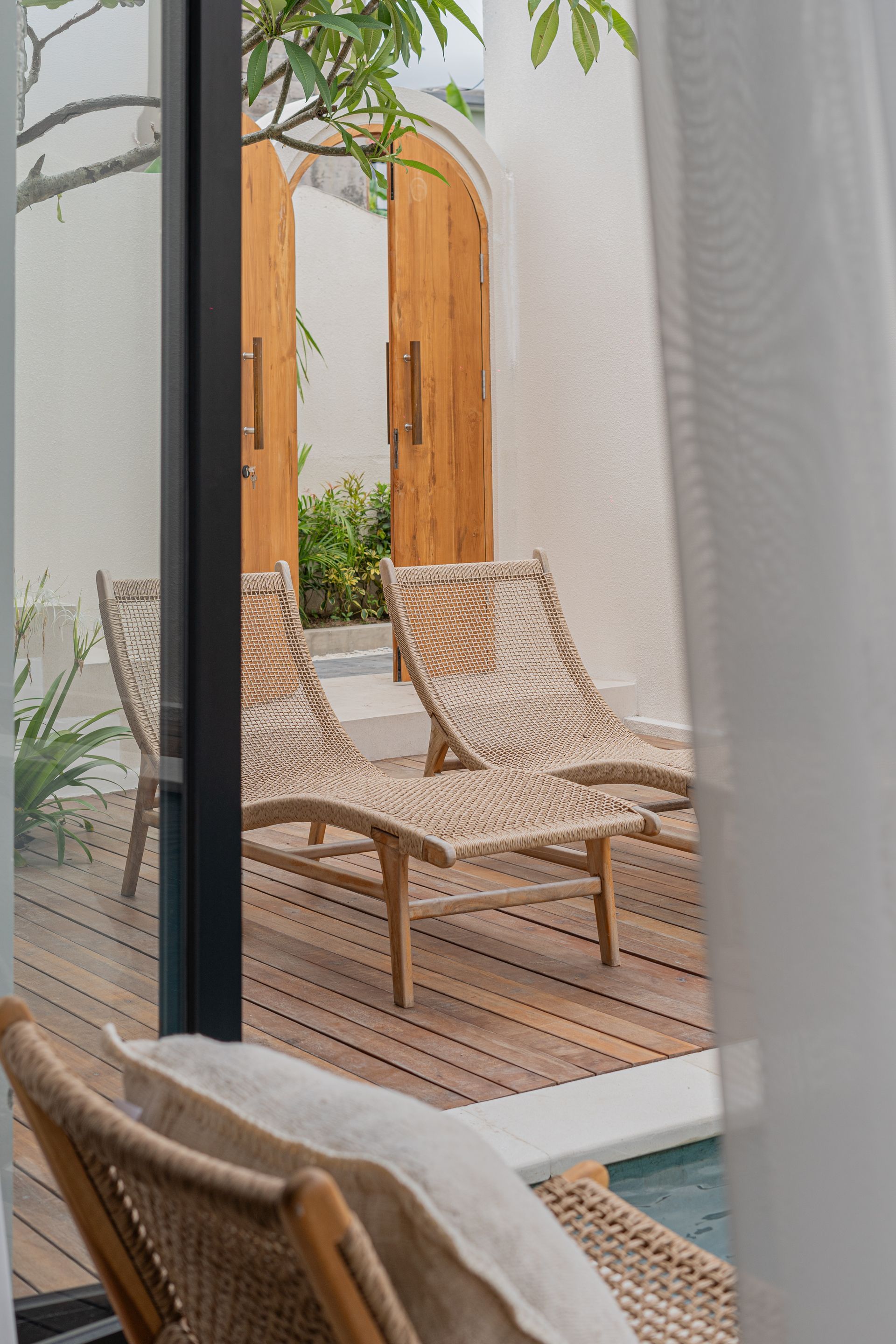Cozy patio with woven lounge chairs, wood decking, and white curtains by a pool, seen through a doorway