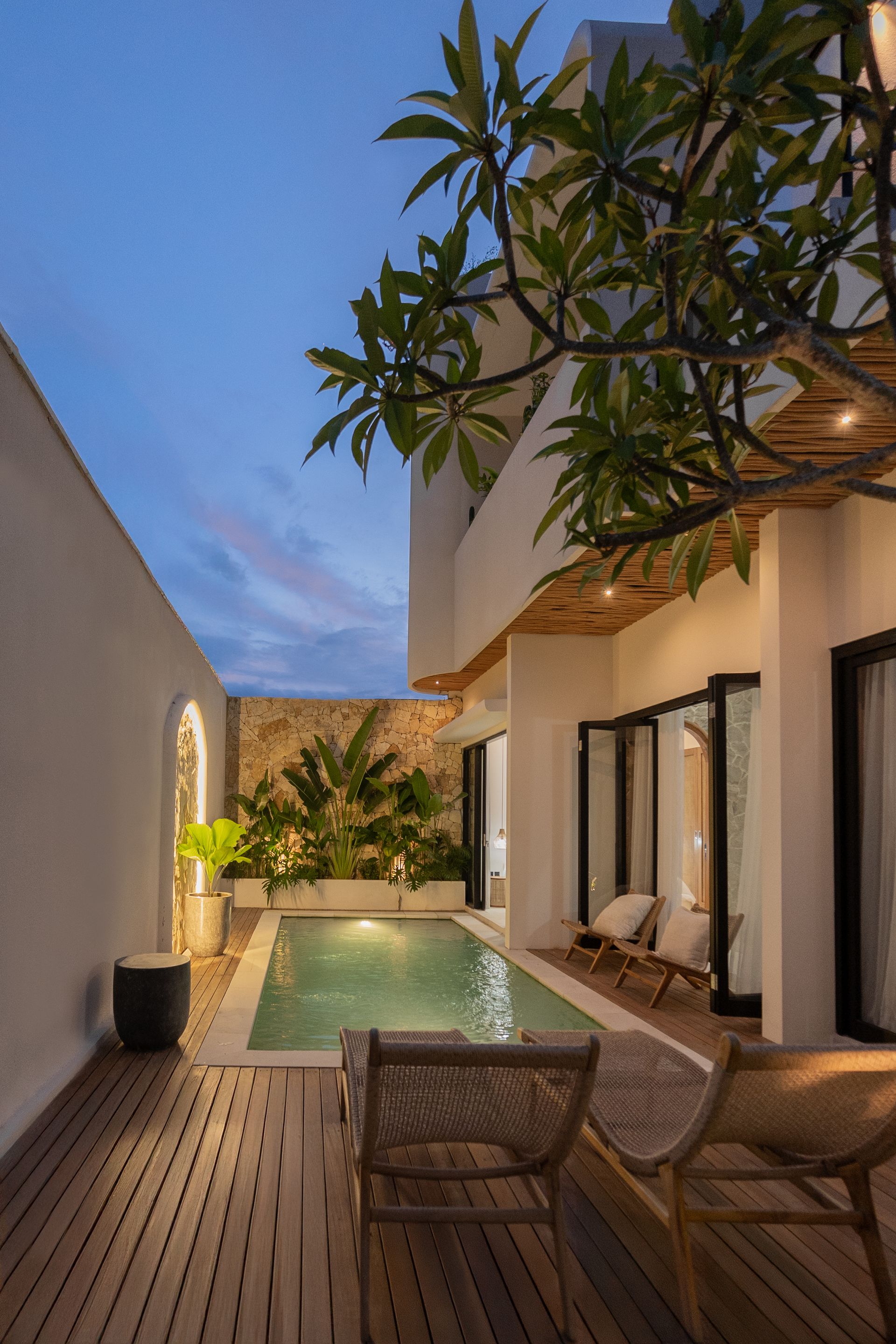 Narrow outdoor patio with a small pool, lounge chairs, wooden deck, and potted trees at dusk