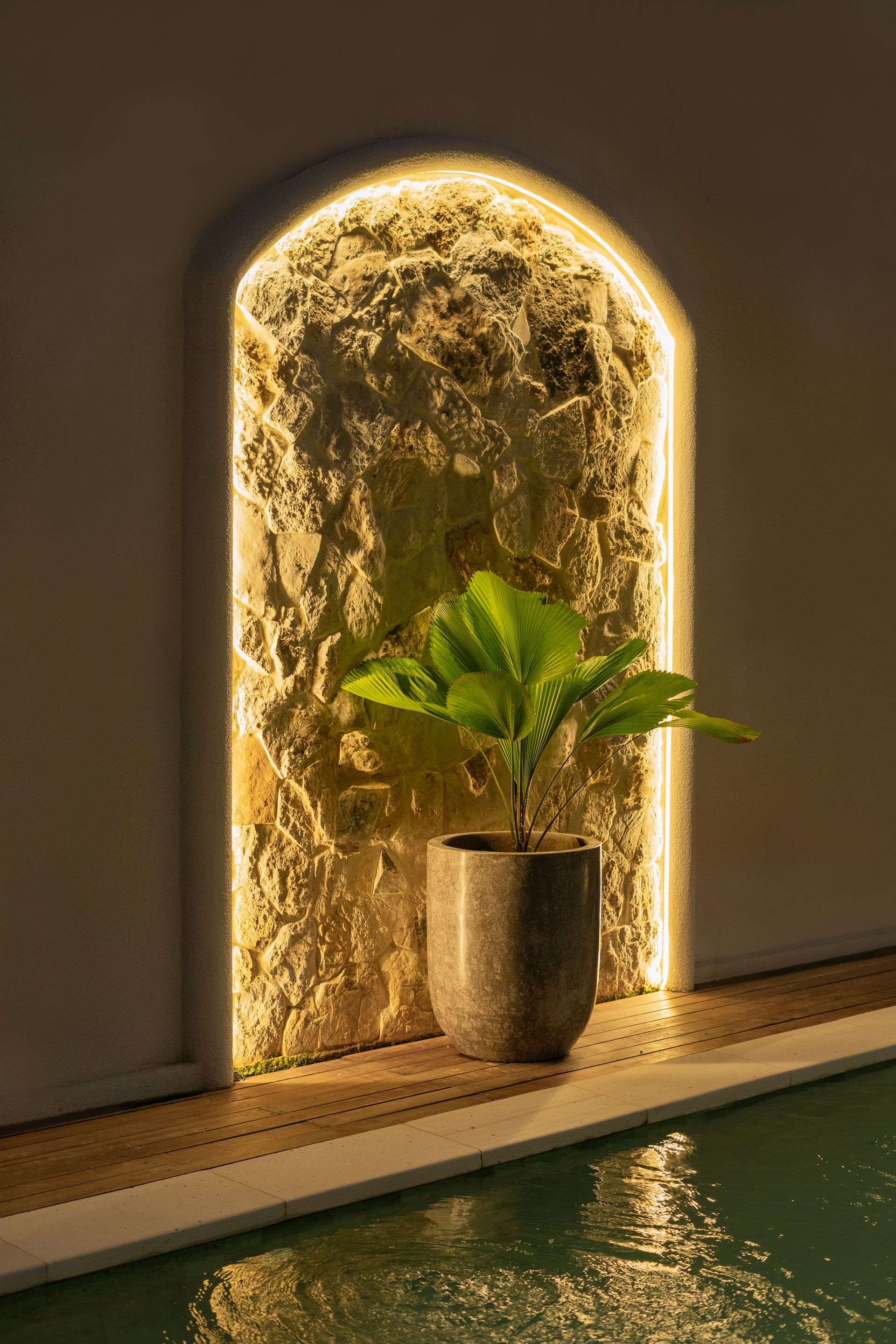 Indoor spa alcove with a potted plant beside a glowing arched waterfall wall and pool edge