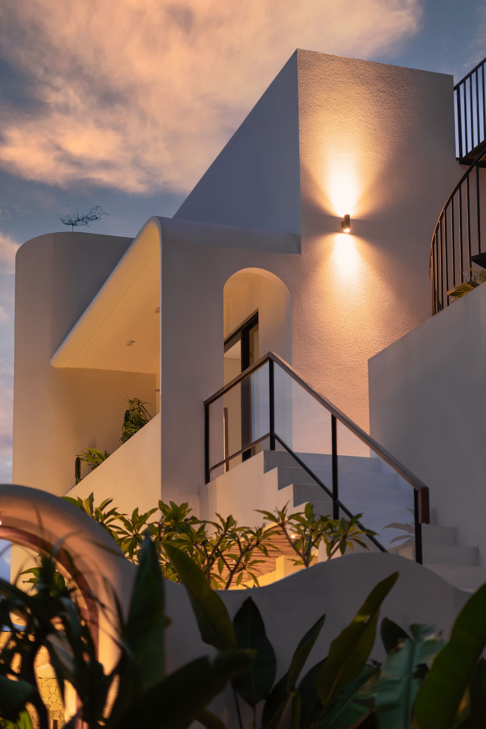 Modern white building exterior at dusk with lit wall lamp, stairs, and plants under a cloudy sky