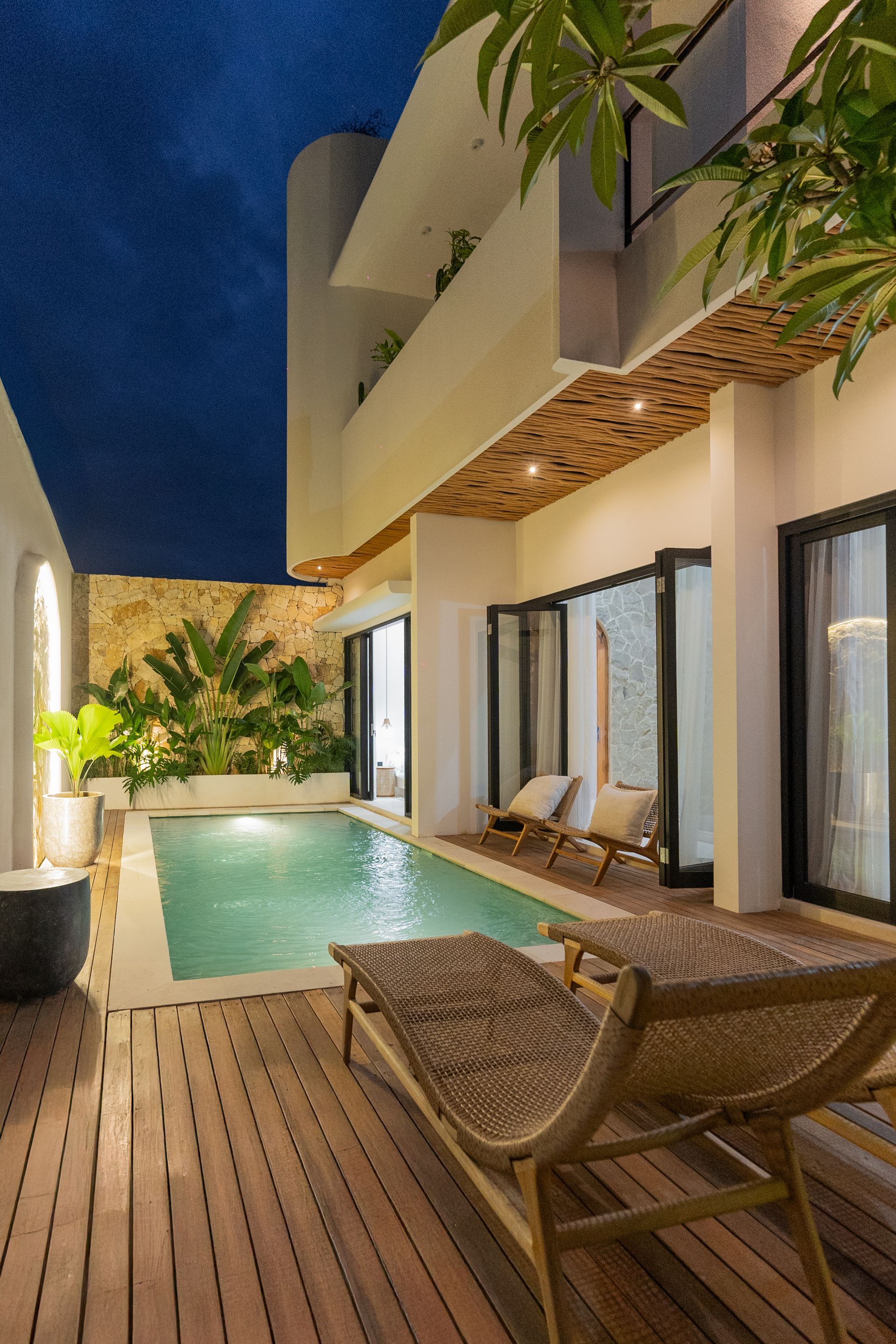 Modern patio with narrow pool, wooden deck, lounge chairs, and lit house exterior at dusk