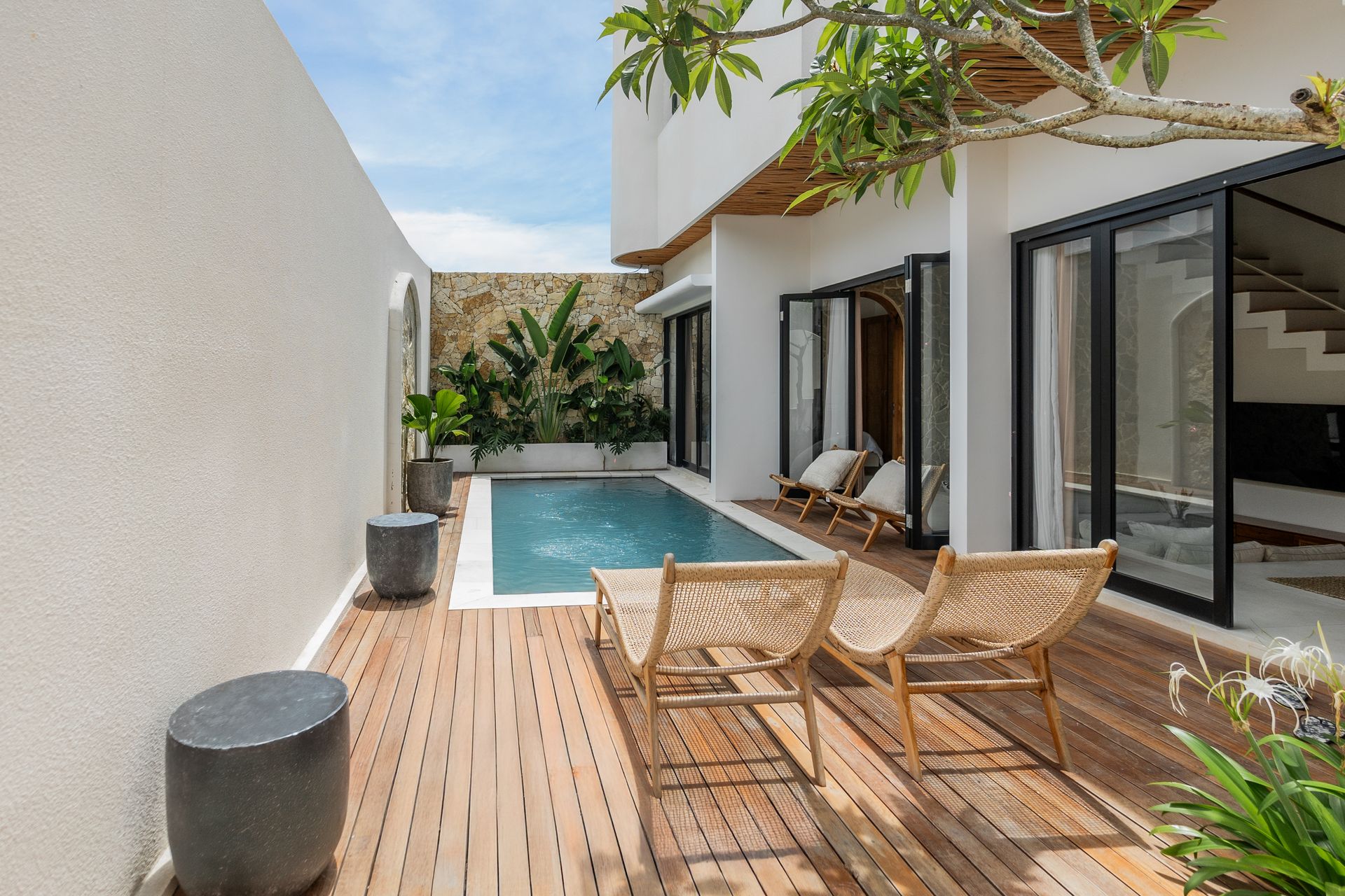 Modern patio with wooden deck, lounge chairs, small pool, and open glass doors beside a white house