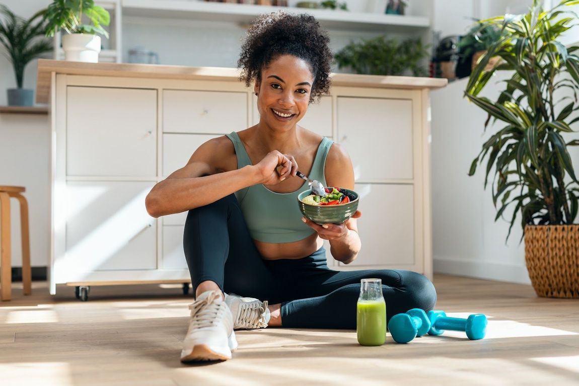 Woman sitting on a sunlit floor eating a healthy salad, with a green smoothie and dumbbells nearby.