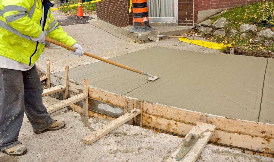 Worker pouring concrete into sidewalk form beside a street under construction