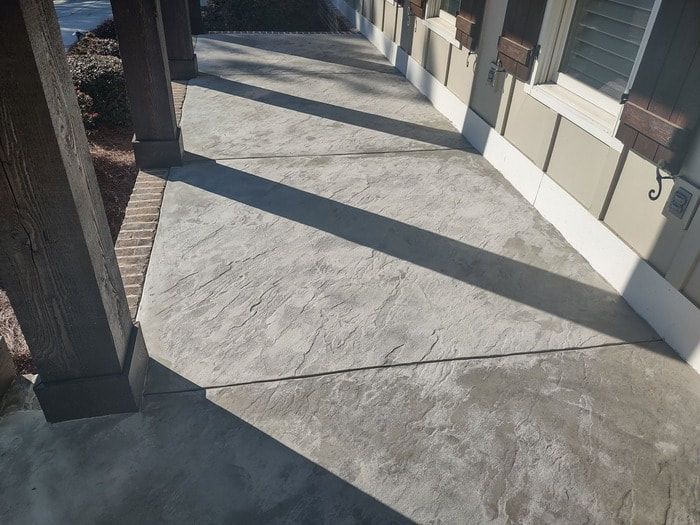 Sunlit concrete sidewalk beside a building with windows and brick columns, casting long shadows.