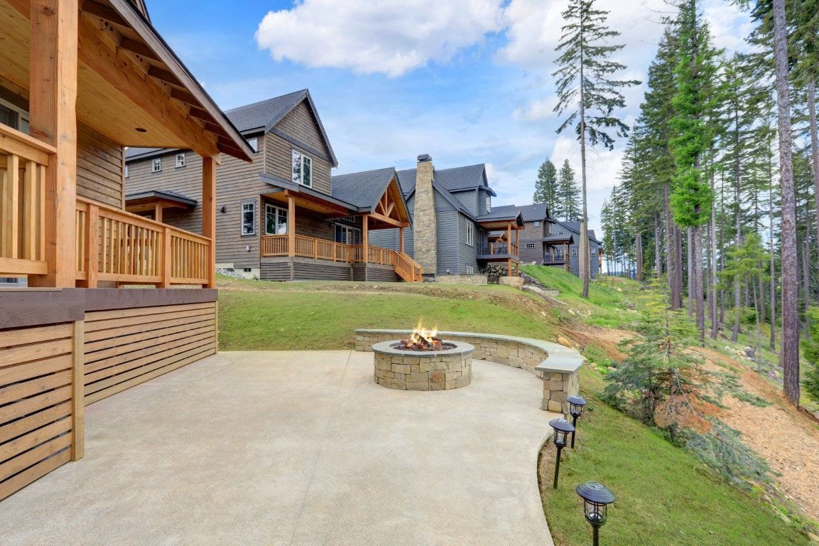 Wooden lodge cabins with decks beside a stone fire pit on a grassy hillside among tall pine trees