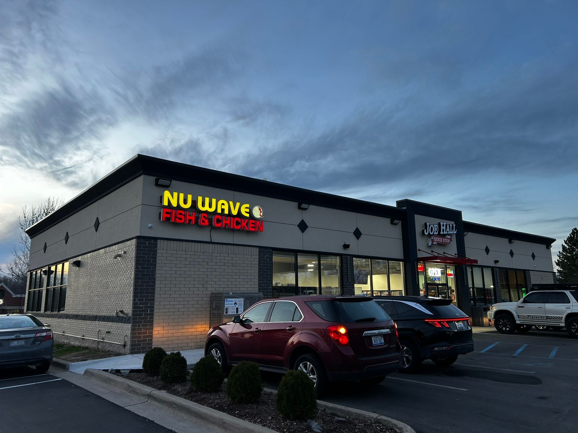 Strip-mall restaurant exterior at dusk, with parked cars and illuminated NU WAVE Fish & Chicken sign.
