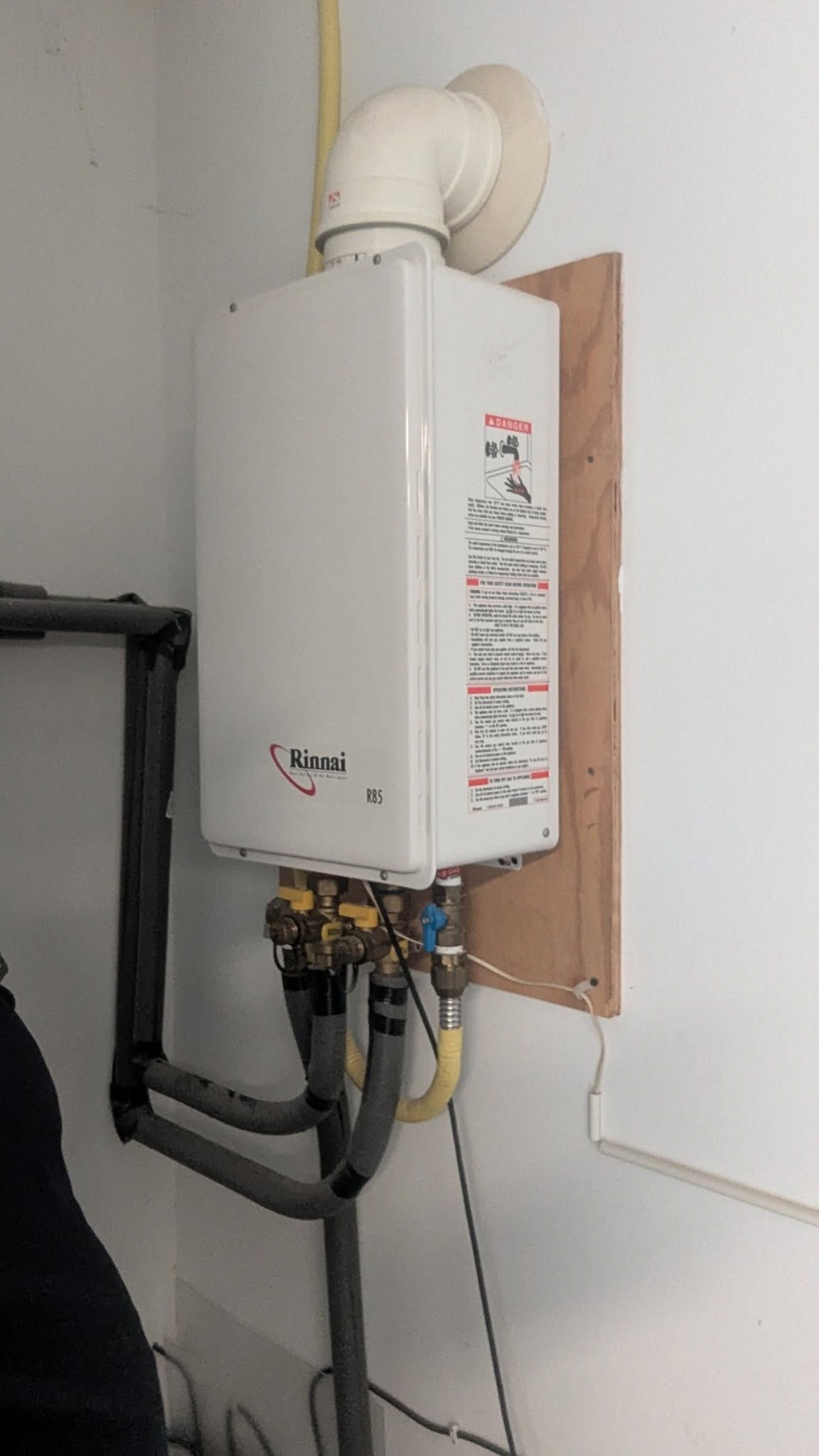 Wall-mounted white gas water heater with vent pipe and exposed hoses in a utility room.