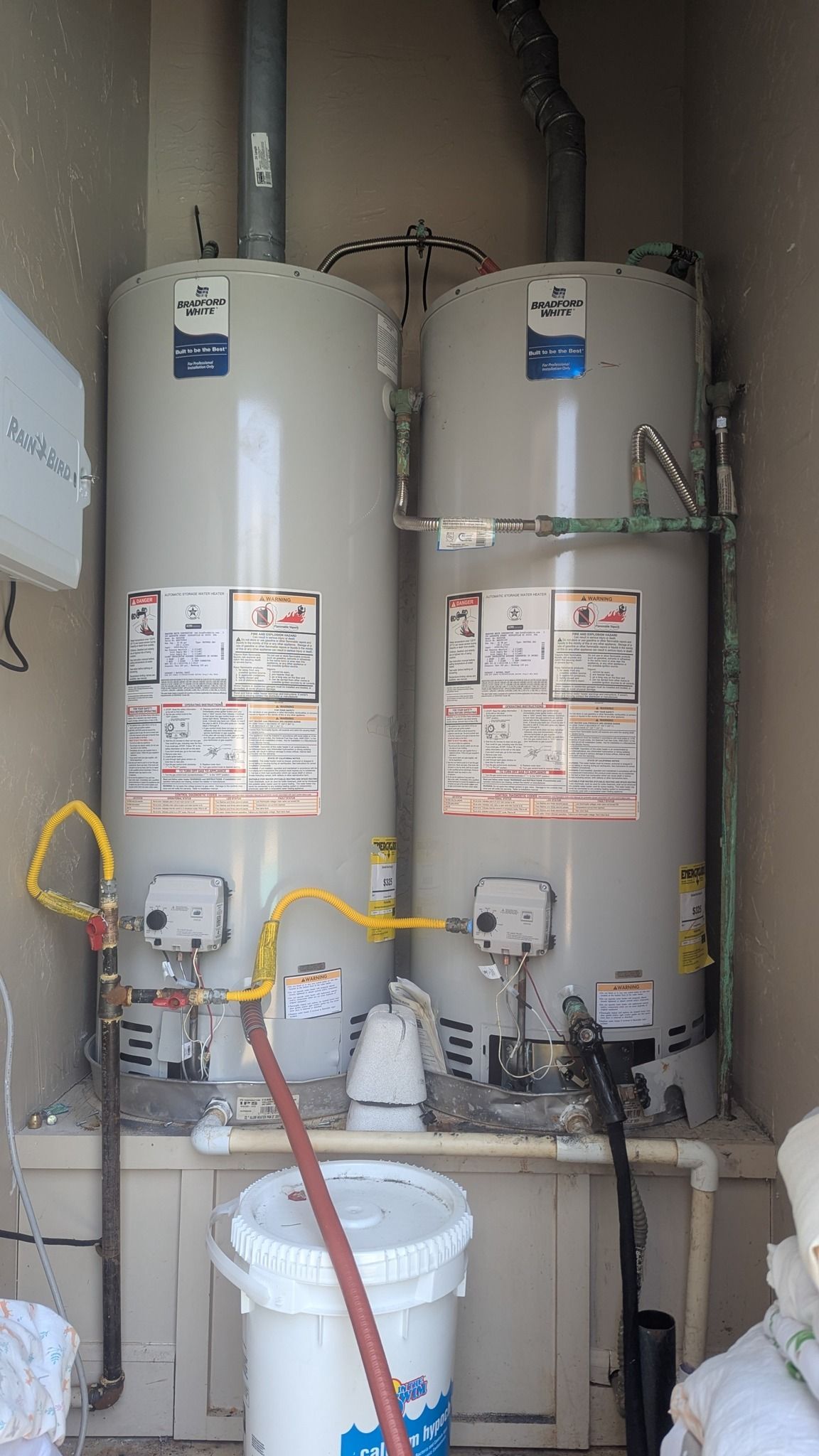 Two tall, silver water heaters side-by-side in a small alcove with pipes and a bucket.