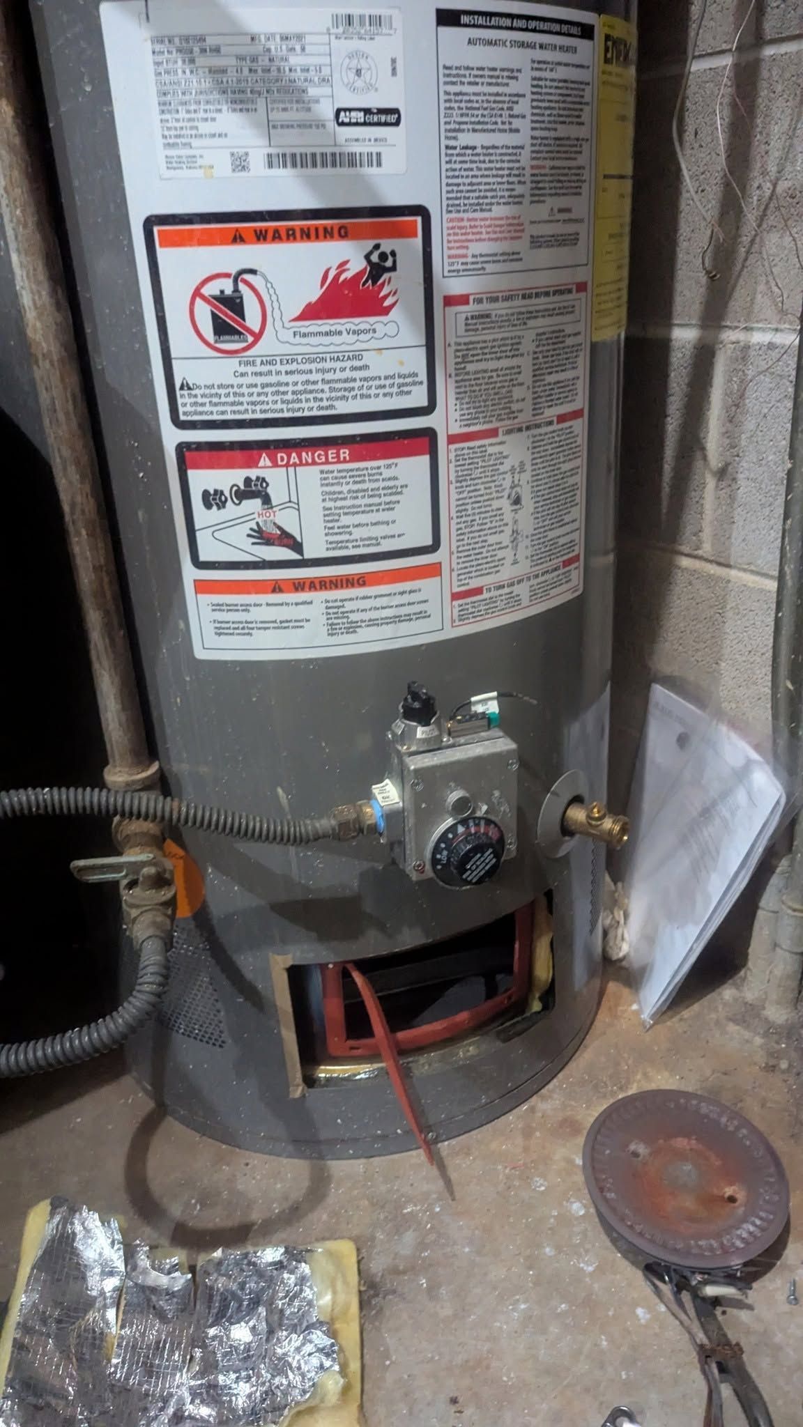 Water heater in a utility closet with warning labels, gas valve, red hose, and a round metal cover on the floor.