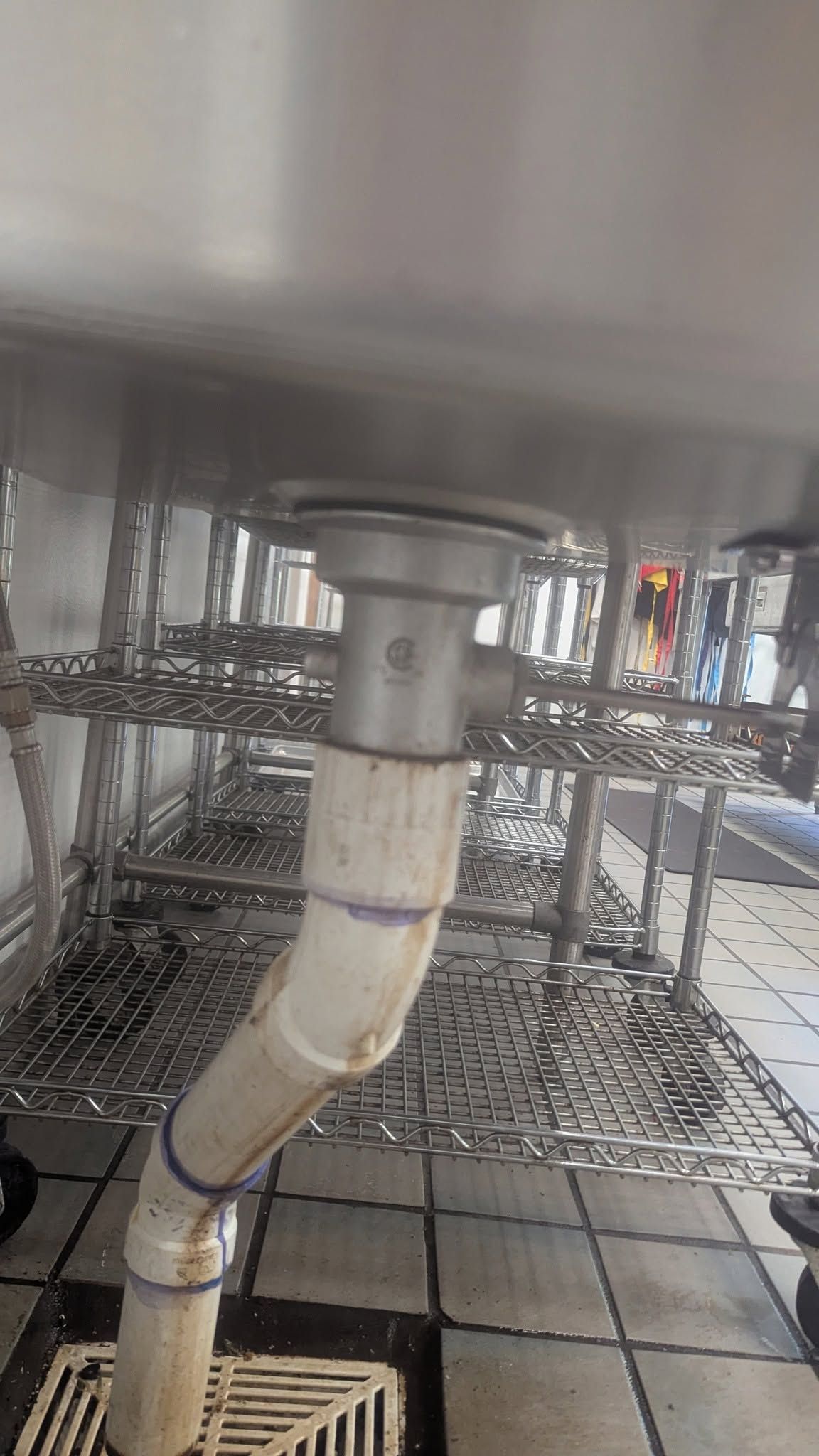 Close-up of a white pipe under a metal sink, with shelving and tiled floor in the background