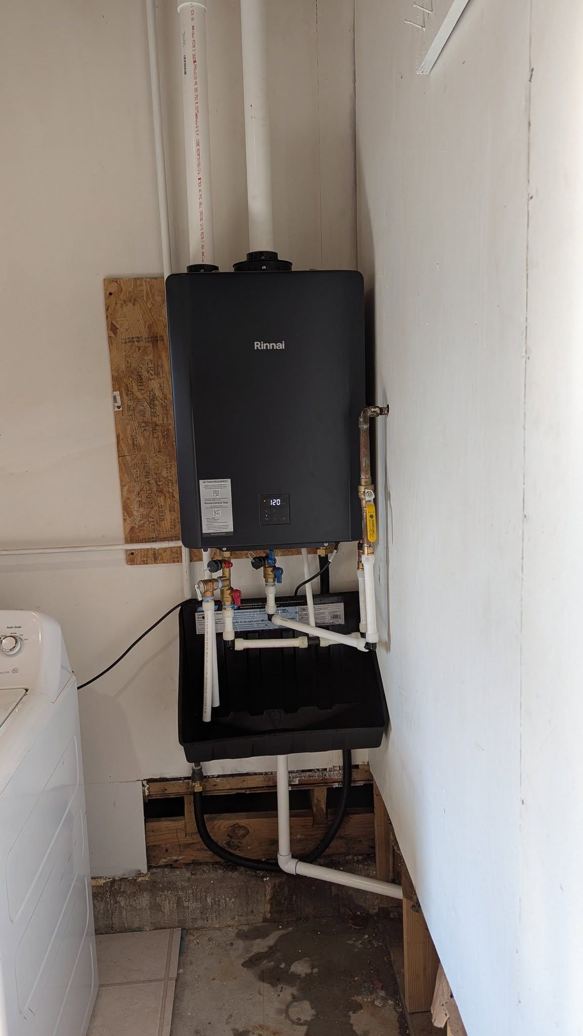 Black wall-mounted water heater in a narrow utility closet with exposed pipes and floor damage