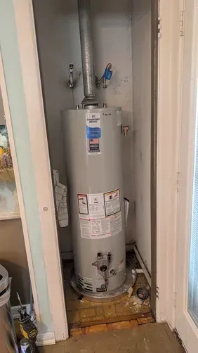Water heater in a narrow utility closet with pipes, valves, and a tiled floor.