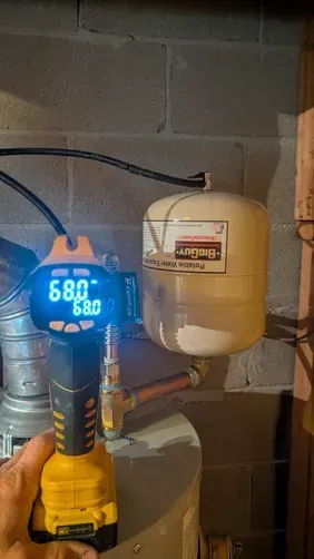 Digital clamp meter on a yellow drill beside a clear jug in a dim utility area with cinder block wall