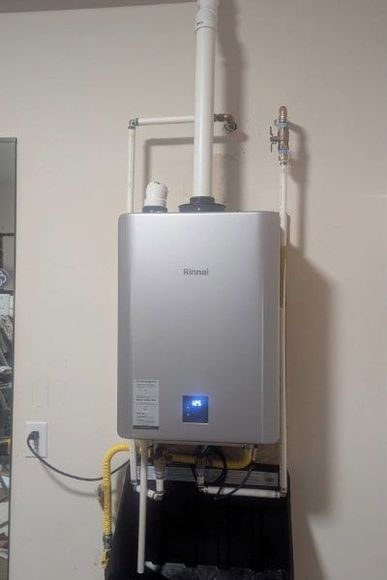 A wall-mounted, silver Rinnai tankless water heater with a white vent pipe and gas line.